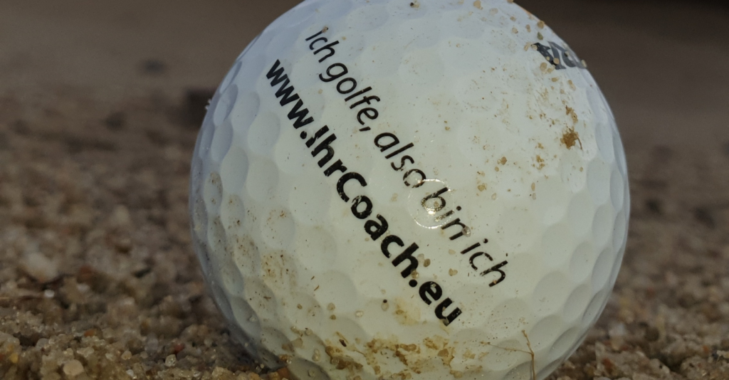 Golfball im Sand mit dem Text Ich golfe, also bin ich